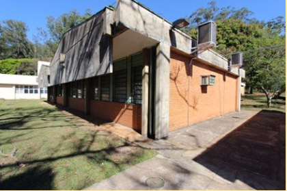 Back and side of the building of the Graduate Program in Special Education. It is day, with sun and blue sky. In the center of the photo there is a building with an external cladding of brick and concrete detail in the corners and roof of the building. Two walls appear in the image. On the wall on the left side there are glass windows and on the side, on the floor, there is grass. On the wall on the right, there are three air condensers at the top. The floor on this side of the building is concrete. At the back of the building there is a forest with trees and a yellow building on the left side of the photo.