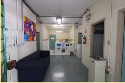 Entrance lobby of the Graduate Program in Special Education building. The walls are beige. On the left side there are two electrical boxes and a colorful painting on the wall. Underneath is a black sofa. In the background is the pantry, an environment with a counter with posters nailed to the front. On the right side is a drinking fountain, an open door and a fire extinguisher on the wall. The ceiling has lighted lamps and the floor is gray vinyl.