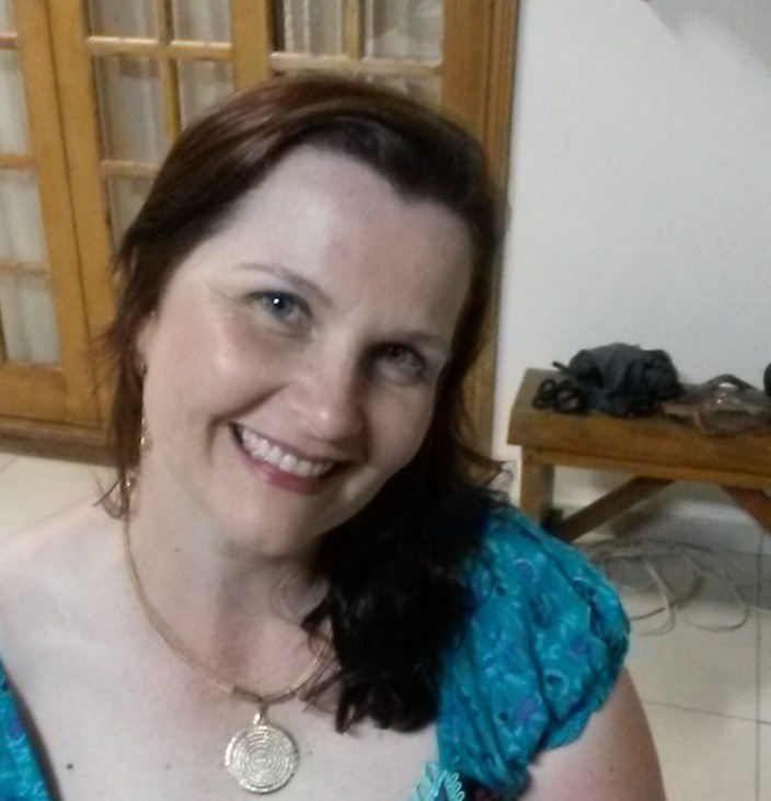 Professora Adriana Garcia Gonçalves. Mulher branca, olhos verdes, cabelo castanho, liso e na altura dos ombros, colocados para o lado e a cabeça esta inclinada para o lado. Ela usa batom, roupa na cor azul, colar dourado com pingente e sorri.