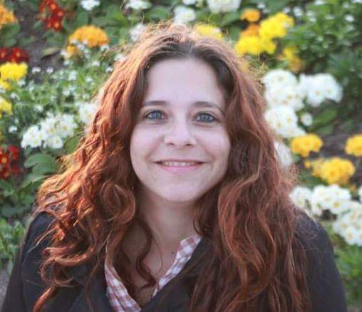 Professora Carla Ariela Rios Vilaronga. Mulher branca, olhos verdes, cabelos ruivos, ondulado, solto e longo. Ela usa batom rosa, camisa xadrez nas cores branca e rosa, e por cima um casaco marrom. Ela sorri. Ao fundo um jardim.
