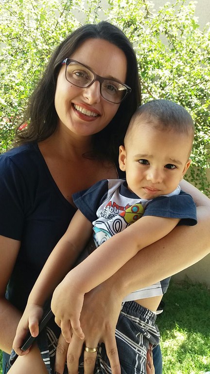 Professora Carolina Severino Lopes da Costa. Mulher branca, olhos castanhos, cabelo presto,liso, solto e na altura dos ombros. Ela usa óculos de grau, batom na cor marrom, camiseta na cor azul marinho, segura uma criança no colo e sorri. Ao fundo plantas verdes.