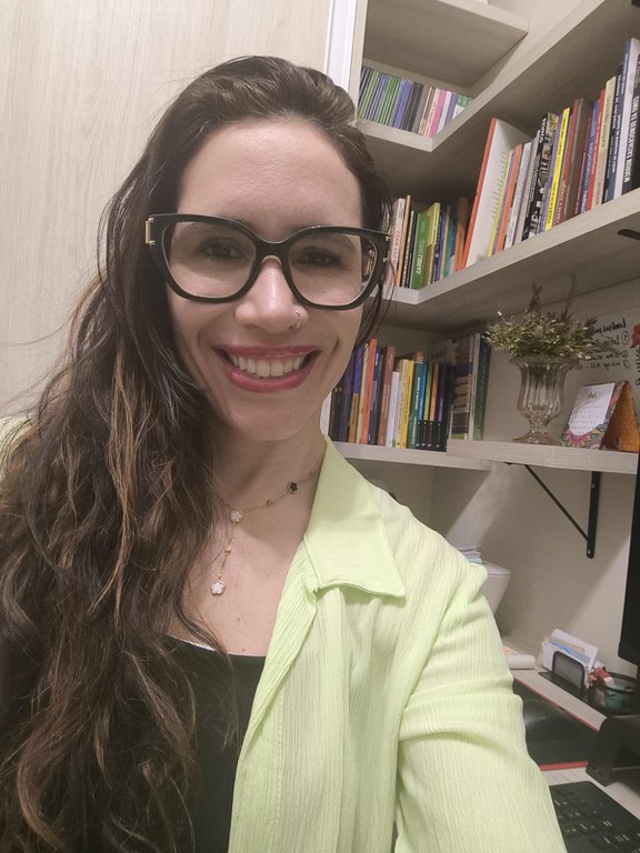 Professora Vanessa Regina de Oliveira Martins. Mulher branca, olhos castanhos, cabelo preto, liso, solto e longo. Ela usa batom rosado, roupa na cor rosa, óculos de grau com armação vermelha e sorri.