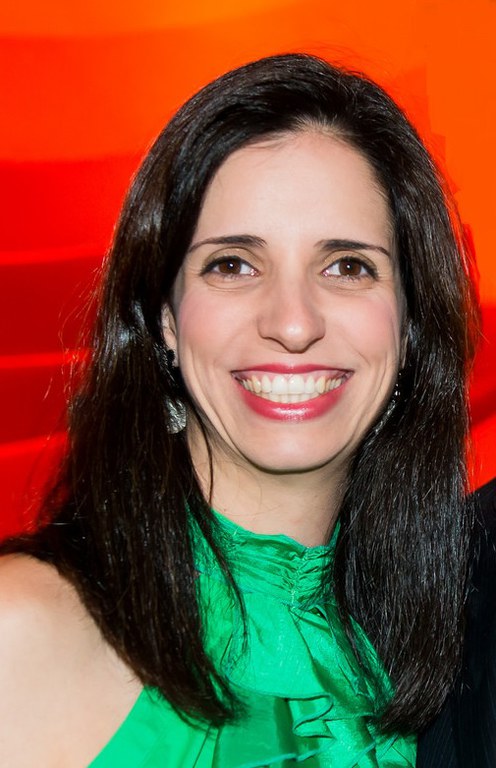 Professora Lara Ferreira dos Santos. Mulher branca, olhos verdes, cabelo castanho, liso, solto e longo. Ela usa batom vermelho, roupa na cor verde e sorri. O fundo da foto é na cor laranja.