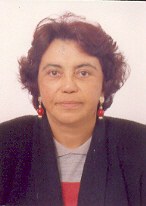 Professora Maria da Piedade Resende da Costa. Mulher parda, olhos castanhos, cabelo castanho, ondulado, solto e curto. Ela usa brinco longo com esfera na cor vermelha, blusa na cor cinza com casaco por cima na cor preta.