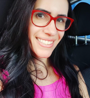 Professora Vanessa Regina de Oliveira Martins. Mulher branca, olhos castanhos, cabelo preto, liso, solto e longo. Ela usa batom rosado, roupa na cor rosa, óculos de grau com armação vermelha e sorri.