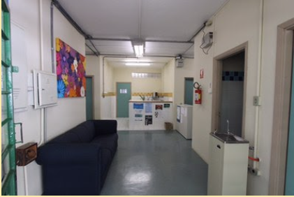 Saguão de entrada do prédio do Programa de Pós-Graduação em Educação Especial. As paredes são na cor bege. Ao lado esquerdo tem duas caixas elétricas e um quadro de pintura colorido na parede. Embaixo tem um sofá de cor preta. Ao fundo está a copa, ambiente com um balcão com cartazes pregados na frente. No lado direito há um bebedouro, uma porta aberta e um extintor de incêndio na parede. No teto tem lâmpadas acesas e o piso é de vinílico na cor cinza.