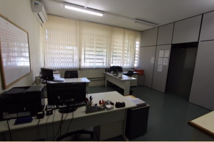 Sala da secretaria do prédio do Programa de Pós-Graduação em Educação Especial. A sala tem três mesas, espalhadas da seguinte maneira: duas ao fundo e uma na frente. Todas as mesas tem uma cadeira e sobre elas há objetos. Na parede do lado esquerdo tem um quadro branco. Ao fundo tem janelas com cortinas persiana vertical. A parede do lado direito tem uma divisória em MDF com porta e cartazes pregados. No teto tem lâmpadas acesas e o piso é de vinílico na cor cinza. traduzir para o espanhol
