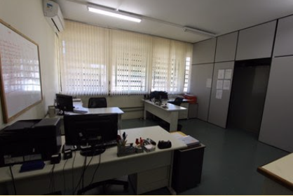 Sala da secretaria do prédio do Programa de Pós-Graduação em Educação Especial. A sala tem três mesas, espalhadas da seguinte maneira: duas ao fundo e uma na frente. Todas as mesas tem uma cadeira e sobre elas há objetos. Na parede do lado esquerdo tem um quadro branco. Ao fundo tem janelas com cortinas persiana vertical. A parede do lado direito tem uma divisória em MDF com porta e cartazes pregados. No teto tem lâmpadas acesas e o piso é de vinílico na cor cinza.